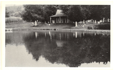 This is a black and white picture of an a shallow, fish-filled pond in front of an old office building. The building is small, with a wrap-around porch, and has a steep pitched roof. 