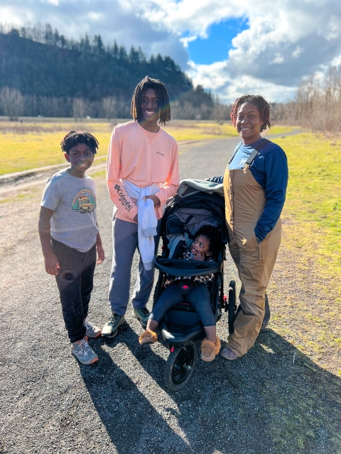 Four family members of different ages, one in a stroller, stand on a gravel road on a sunny day with trees and clouds in the distance