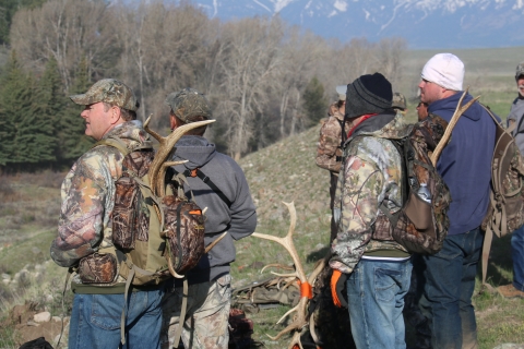 Men in hunting jackets and caps or knit hats stand on hillside with antlers protruding from their backpacks.s