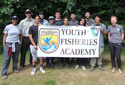 A group of people stand together outdoors holding a large banner that says "Youth Fisheries Academy" with an FWS logo.