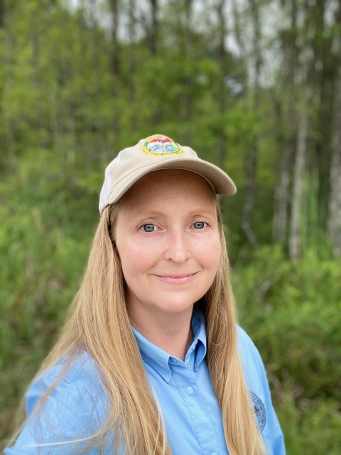 A woman smiles at the camera. She has long, blonde hair and is wearing a baseball cap. Her button up shirt is blue. She is standing in a green forest. 