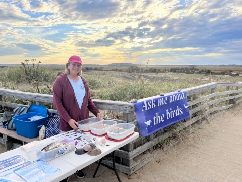 a woman wearing a red ball cap and maroon sweatshirt stands behind a table with information, with a beach and cloudy sky in the background