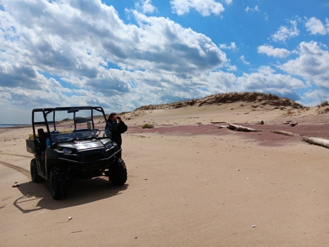 a woman stands beside a utility vehicle on a beach looking through binoculars, with a sand dune and clouds and blue sky in the background