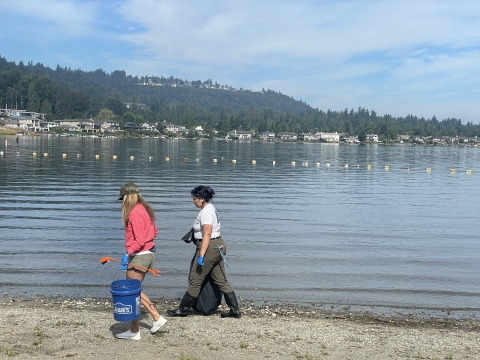 Two people walk with litter-collecting tools in front of a lake