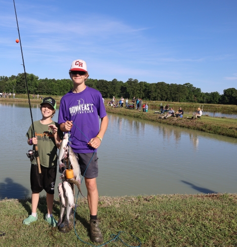 2 Youths, one holding catfish stringer and the other holding a fishing rod and reel in front of pond.