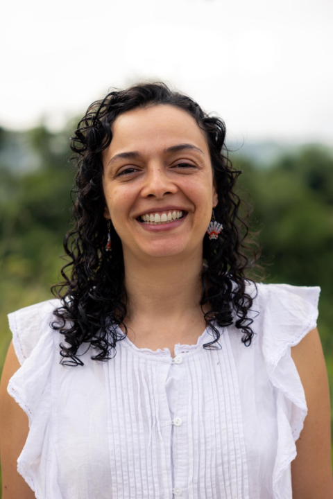 A young Hispanic woman smiles directly into the camera. She's wearing a white blouse, colorful earrings, and her brown hair is down and curly. 