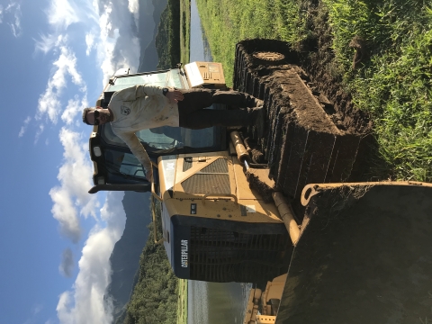 A man in a dirty brown uniform stands on a tractor in a wetlands landscape surrounded by dramatic mountain ridges. 