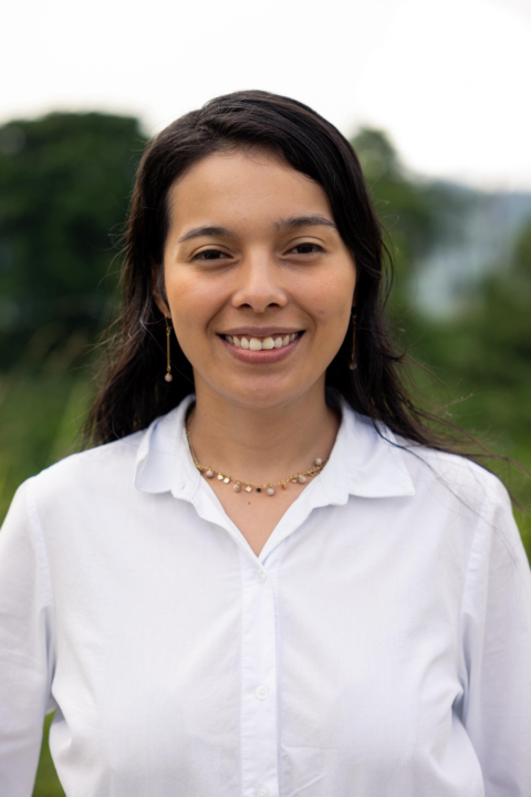 A young Hispanic woman smiles into the camera. She's wearing a white button-up shirt, long drop earrings and a studded necklace with gold tones, and dark brown hair that's down behind her shoulders. 