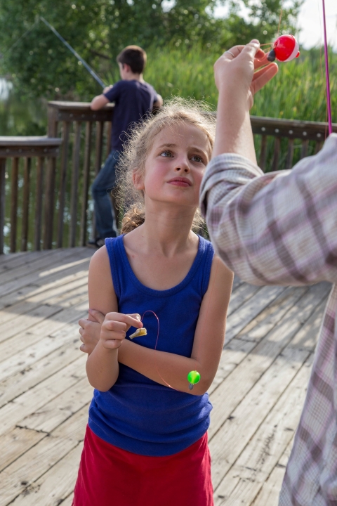 A girl in a blue shirt and red skirt stands before an adult who is attaching a bobber to a fishing pole. They are standing on a fishing pier, with another angler in the background.