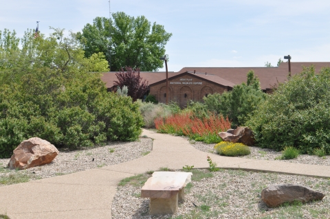 Shrubs and red and yellow flowers line a path leading towards a brick building labeled "Deer Flat National Wildlife Refuge."