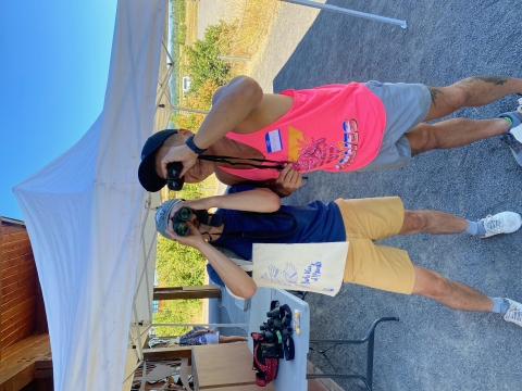 Two folks in brightly colored clothes look through binoculars. 
