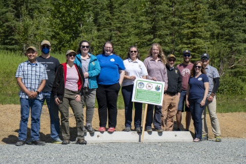 Eleven people pose together for a picture at the edge of a gravel parking lot with a sign with a wooded background. The sign has logos of the Alaska Department of Fish and Game, Wildlife Restoration, and Alaska State Parks on it. It reads: Eklutna Lake ATV Trailhead Chgach State Park Expansion of this parking lot was provided through a cooperative effort between the Alaska Department of Fish and Game, Alaska State Parks, and the U.S. Fish and Wildlife Service Wildlife Restoration Program. 