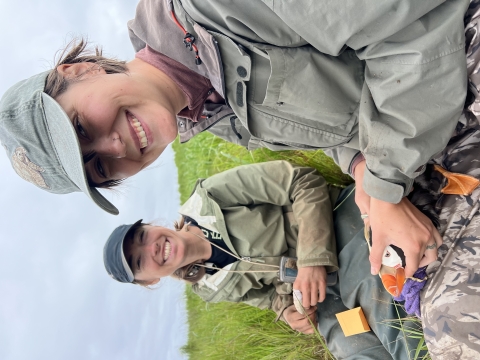 Two women sit on a hillside and one is holding a tufted puffin.