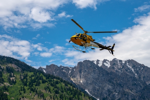 A small yellow helicopter with a crew member looking out soars above rocky mountains in Wyoming.