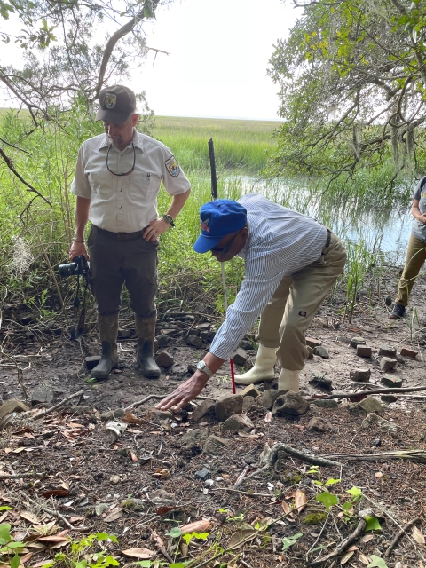 Two men, one standing and one bending and touching the earth, examine the site where a house once stood.