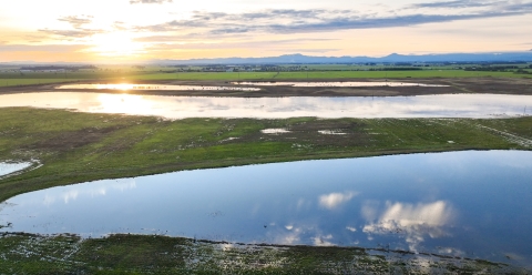 Wetland scene from the air, with clouds reflected in water and hills and sun in the distance.