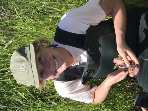 A woman wearing overalls and a hat, smiling at the camera while holding a small salamander in her hands