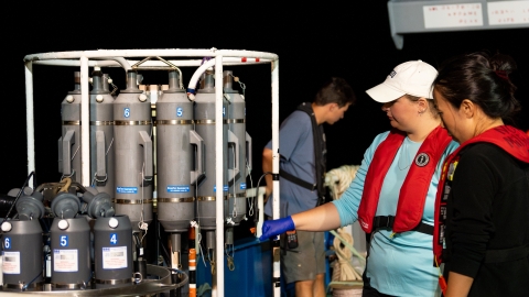 An Asian female scientist, and a Caucasian female graduate student are looking at the equipment on the deck of the ship. A young, male Caucasian crew member is working in the background. It is pitch black. All people are wearing personal flotation devices.