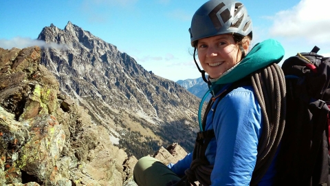 A woman wearing a backpack, rope, and climbing helmet sits high up with a mountain view in the distance