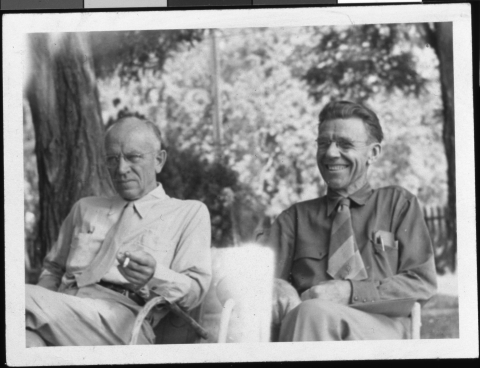 Close-up of two men (Aldo Leopold and Olaus Murie) sitting alongside each other in metal lawn chairs. Both wear shirts with ties. Leopold looks serious, while Murie has a wide smile. A few trees are in the background, and a picket fence can just been seen.