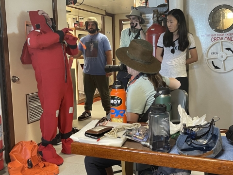 Four members of the expedition team are looking on as the Captain is zipping up a bright red suit that protects people from the elements should they need to abandon ship. They are inside the ship and there is lots of gear on the table in the foreground.