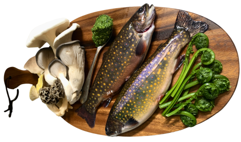A wood platter with various mushrooms, fiddlehead ferns, two fish with gold dotted and green-brown scales, and a spoonful of green pesto.