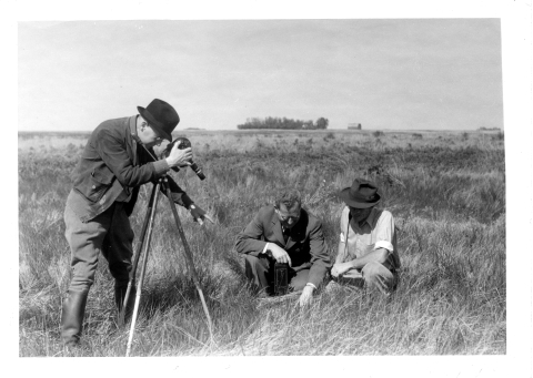 Two men kneeling in the prairie grass, examining a Grouse nest (the nest is not really visible); on the left, and man is operating a camera of 1930's vintage that is mounted on a tripod