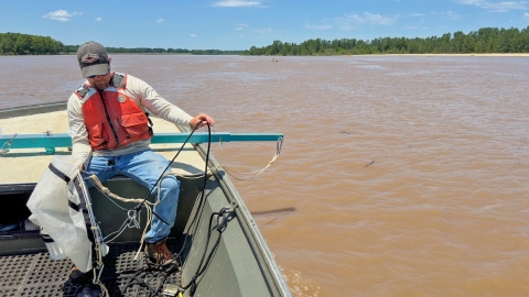 Man sitting on a boat preparing a gill net to deploy in a brown colored river with trees in the background.