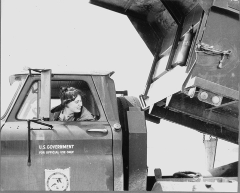 close-up of the driver's compartment of a dump truck, with a woman sitting in the driver's seat, leaning out the open window, and looking back at the raised flatbed of the dump truck. The door of the truck features the logo "U.S. GOVERNMENT FOR OFFICIAL USE ONLY", as well as the Bureau of Sport Fisheries and Wildlife shield