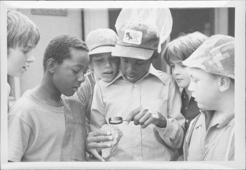 Close-up of a goup of six children (all appear to be boys), gathered around the boy in center who is examining a small rock with a magnifying glass. One of the boys is carrying a butterfly net.