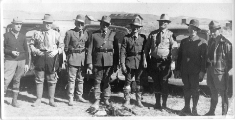 Eight Law Enforcement Officers/Agents posing for the camera, standing in a straight line. Three officers in the center wear Law Enforcement uniforms (consisting of pants, shirt with necktie, coat, and broadbrimmed hat. Several of the other officers wear different brimmed hats (campaign-style hats, fedoras, etc.). In a small pile in front of them is what appears to be several dead waterfowl. Three automobiles of the 1930's vintage are immediately behind them. In the background are buildings and mountain