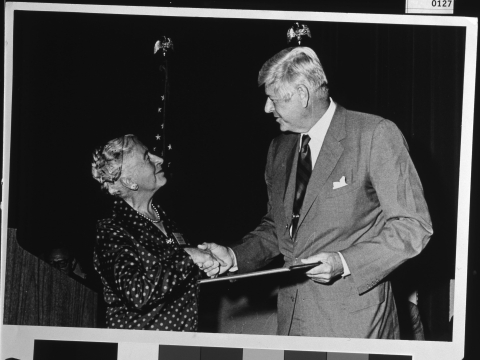 An older woman (Mardy Murie) on the left, shaking hands with an older man (Rogers C. B. Morton) on the left, as they make eye contact. The man (Morton) is holding a plaque in his other hand. Two flags (one of the U. S. National flag) on flag staffs are behind them.