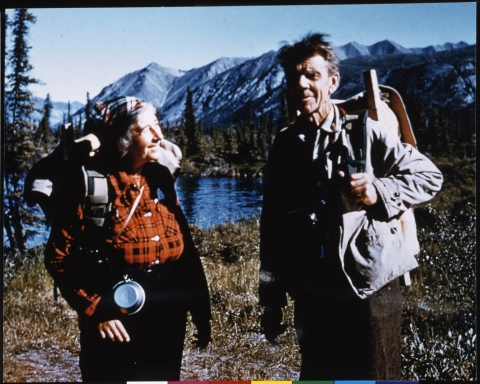 Two people, Mardy and Olaus Murie, standing next to each other, outfitted for a hike, wearing field clothing, and carrying hiking backpacks. In the middle background is a body of water (the Sheenjek River), with a mountain line in the far background