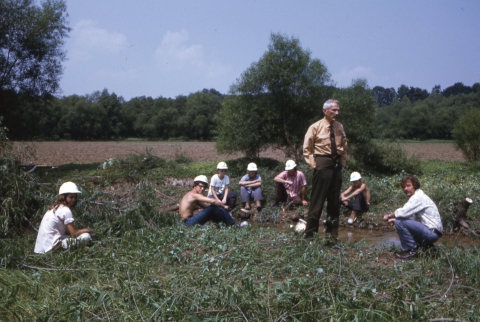 An older man (Refuge Manager Tom Atkeson), in U.S. Fish and Wildlife Service uniform of the early 1970s, standing near a small tree in a clearing; seated around him on the ground are about 6 young people (Youth Conservation Corp members), all of whole are wearing hard hats. An adult (without a hat) sits kneels near Atkeson