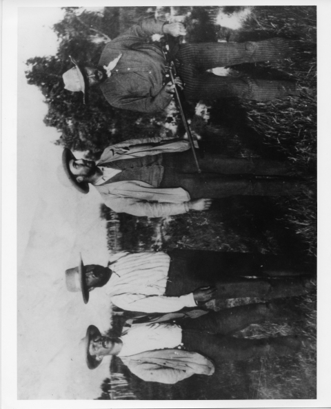 Four men posing together for a photograph; all of them wear 1890's vintage field clothing, including broad-brimmed hats (it makes them looks like cowboys). The man on the far right (Bureau of Biological Survey manager C. Hart Merriam) is holding a shotgun in from of him, level with his waist. A few small trees are in the background.