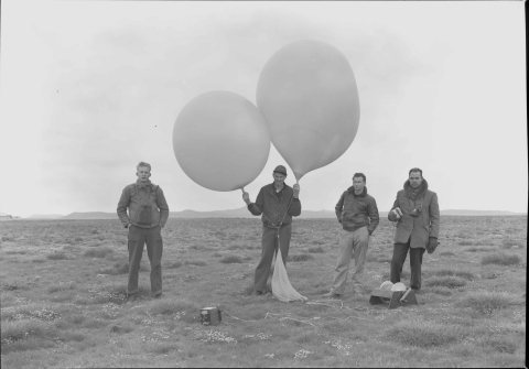 Four men standing in a featureless area, with low grass; and a mountain line in the background. The man in the center holds two large meteorological balloons, to which is attached a bag, with a rope line or cord leading to a winch that is placed on the ground, in front of the man on the far right. A camera of 1940s vintage lays in the ground in the foreground, also attached to a length of rope/cord. All four men are dressed from warmth.