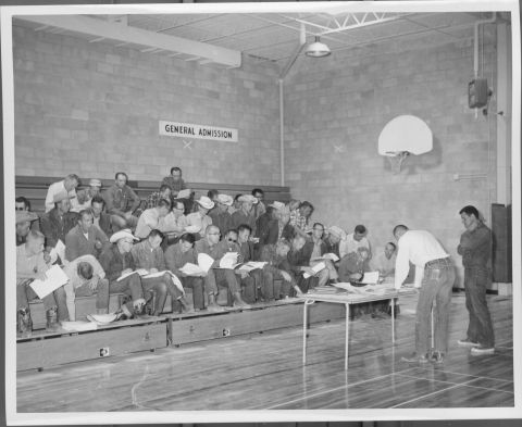Around 36 men sitting on bleachers in a high school gymnasium, with many examining/reading the sheets of papers they have apparently been given. In front of them, on the gym floor, is a small table, with one man leaning over it, examining something one the table surface, and another man standing just behind him.