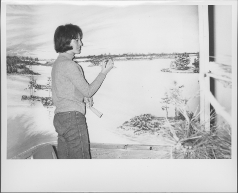 A woman, standing, painting a landscape scene that features fields and patches of trees. She holds a paint brush in her right hand, and what appears to be a cloth in her left hand. A wooden framework appears on the right-hand side, along with what looks to be a pine boughs.