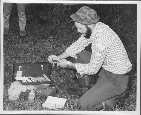 Man kneeling down, holding a small bird (Indigo Bunting) in one hand, while he secures a metal band around the bird's leg. He is kneeling in a grassy area; in front of him is a medium-sized metal case containing bird banding equipment.