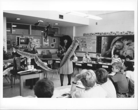 Two teachers, both female, holding up a snake (specifically Anaconda) skin in front of an elementary school classroom, and students sitting in desks in the foreground, and in front of the back wall, center. Clothes and hair styles of 1990s vintage