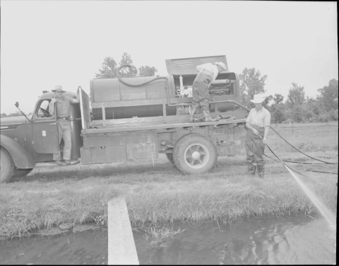 A large tuck of 1940's vintage, with pumping equipment on the flatbed. One man stands on the right, at the back of the truck (the truck is parked so that the side is facing the camera), and is spraying liquid from a hose that is attached to the equipment on the truck; he is spraying it in to a channel of water in the foreground. Another man is standing on the flatbed, checking the motor of the equipment. A third man is standing on the running board of cab of the truck.