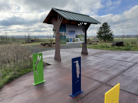 Yellow, blue, and green bike racks in front of an interpretive kiosk. Bike racks have cutouts of a Great Blue Heron and Dragonfly.