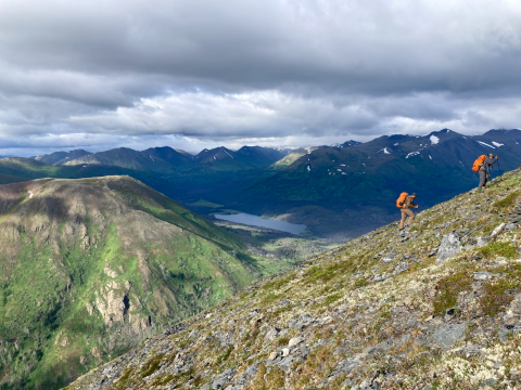 In the foreground, 2 people with large orange backpacks hike up a steep slope of rock and lichen. In the background are big mountains with snow patches on top, a lake in a valley, and a cloudy sky.