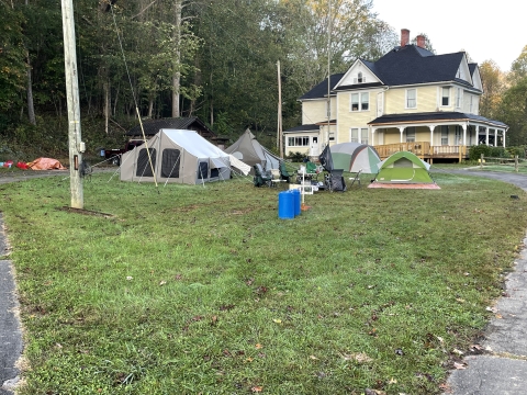 Looking across a green lawn with trees in the background is a large yellow house with a wrap around porch. In the foreground, near the house are several tents set up providing a place for storm volunteers to stay. 