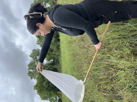 Aaliyah outside holding an insect net. green, long grass and trees in the background