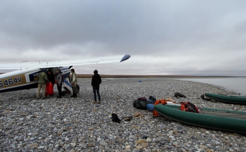 a small group of people by a bush plane and 2 rafts on a gravel bar