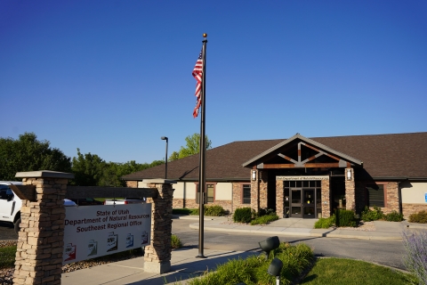 Photographed is an entrance sign next to an American flag on a flagpole. The Sign reads "State of Utah Department of Natural Resources Southeast Regional Office". Behind both is the regional office building. 