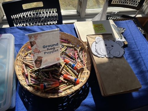 A basket of crayons, a cutout of a cartoon groundhog bag, and a stack of brown paper bags on a table.