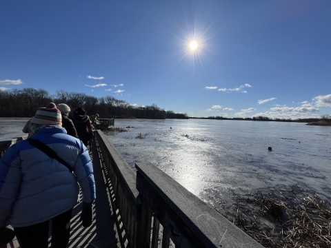 Sun shining over people walking on a boardwalk suspended over a frozen body of water.