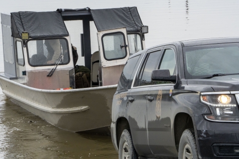 An SUV with a trailer with boat on it is backed into a river boat launch with a Federal Wildlife Officer at the helm. 